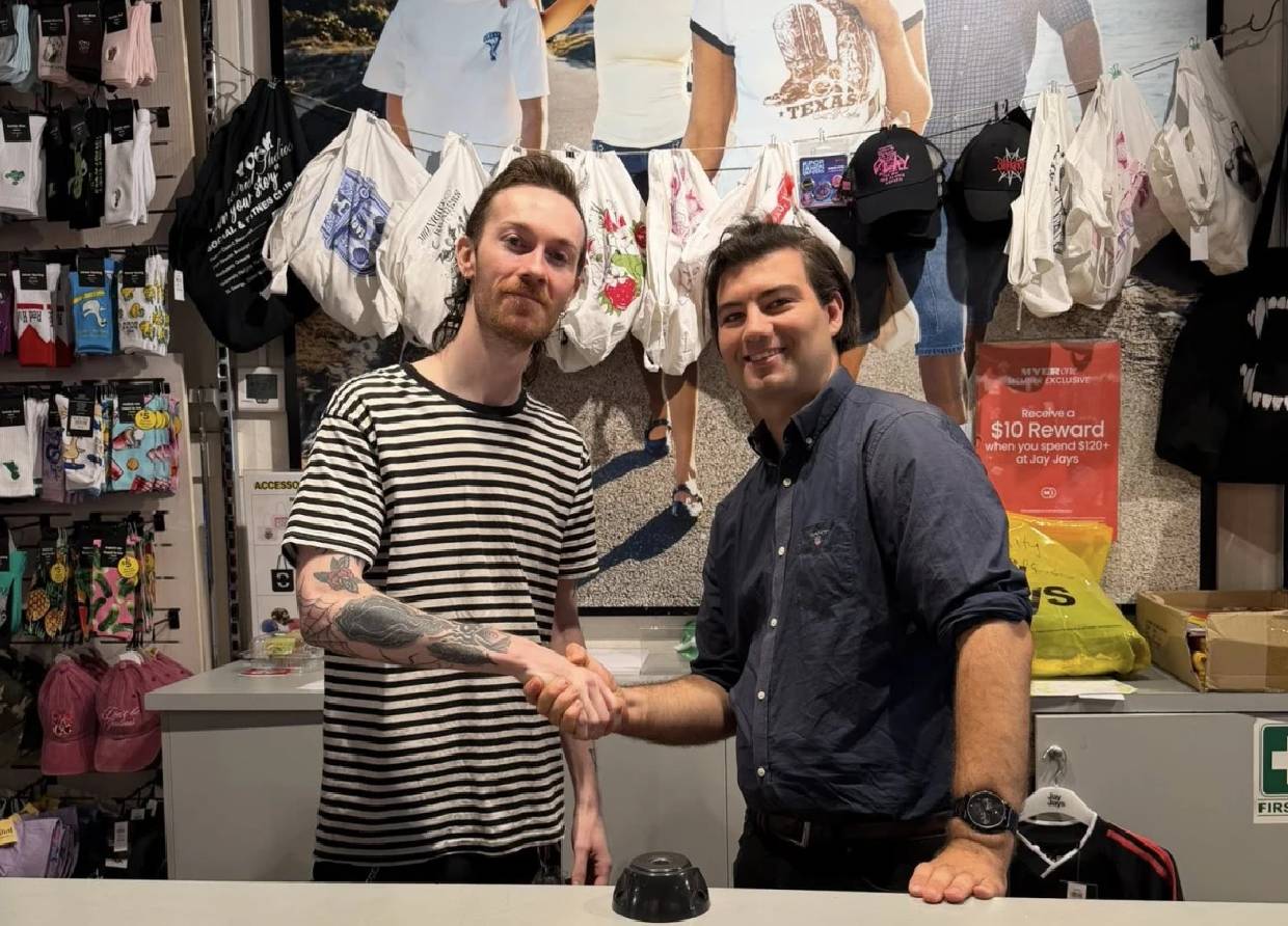 Two men smiling and shaking hands across a counter in a retail store